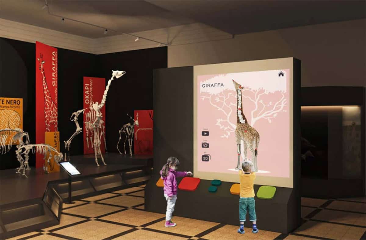 Bambini interagiscono con una giraffa su un grande schermo interattivo al Museo Civico di Zoologia di Roma.